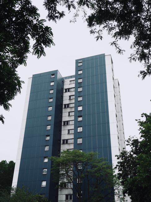 A tall modern residential building with a predominantly blue glass facade and white structural elements, viewed from the ground looking upwards. The building has multiple windows arranged in vertical rows, with some windows open or visible as small dark rectangles. Surrounding the structure are leafy green trees, partially obscuring the lower floors and providing a natural contrast to the urban architecture. The sky above is overcast with grey clouds, and the overall scene suggests a city environment suitable for house removals or home relocation services, as offered by Man With a Van Abbey Wood.