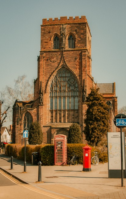 Photograph of a large historic brick church with tall, arched stained-glass windows and a crenellated tower, situated on a street with clear pavement. In front of the church, there are typical UK street elements including a blue cycle lane sign, a red telephone booth, a red post box, and a bicycle parked near a bus stop sign that displays maps and information about Abbey Foregate. The scene is well-lit with natural daylight, and the environment includes some bushes and a few trees nearby. This setting reflects a typical UK town centre, possibly during a mild autumn day, with no visible moving activity or furniture, but the scene provides context for local logistics and transportation services related to house removals and home relocation, as offered by Man With a Van Abbey Wood.