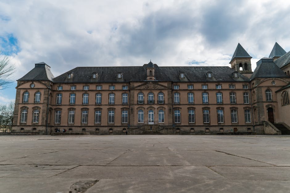 A large, historic building with a symmetrical façade featuring multiple tall, arched and rectangular windows, situated behind a wide paved area. The structure has a dark, shingled roof with several small dormer windows and two prominent towers with steep, pointed roofs on the right side. The exterior walls are constructed from beige or light brown stone or brick. The sky above is partly cloudy with patches of blue. In the foreground, the paved surface is uneven with visible cracks, and three people are seated on the left side near the building. This scene illustrates the exterior of a stately property suitable for a home relocation or furniture transport service, with the building's grand architecture providing context for moving or packing activities managed by companies like Man With a Van Abbey Wood.