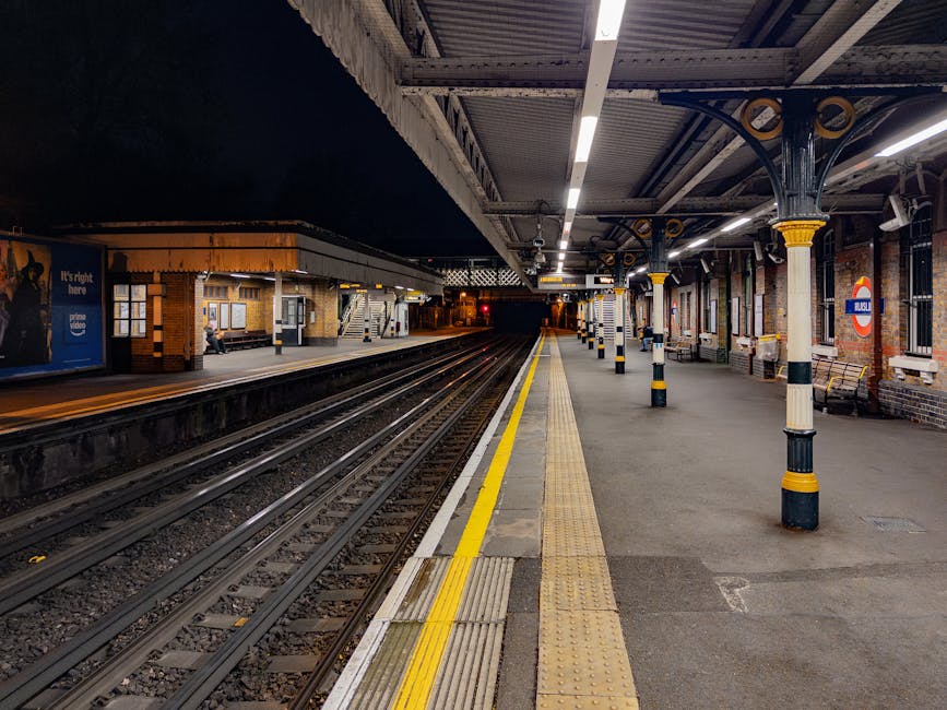A deserted platform at Abbey Wood railway station during evening or night, with the station illuminated by overhead lighting. The platform has a yellow safety line along the edge and tactile paving for visually impaired passengers. Multiple black and yellow striped bollards line the platform, supporting the metal canopy overhead, which features decorative supporting columns painted with yellow accents. On the left, there is a small brick building with windows and a station sign, and a person is sitting on a bench near the building. Parallel railway tracks extend into the distance, disappearing into the dark tunnel beyond the platform. The scene is clear and well-lit, with no moving vehicles or people visible, creating a calm atmosphere suitable for discussing station logistics related to house removals and furniture transport, as part of a home relocation process with Man With a Van Abbey Wood.