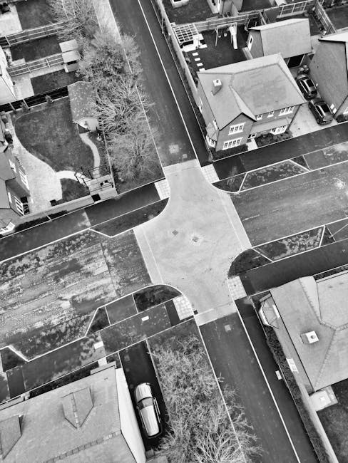 A black and white aerial photograph of a residential area showing a four-way intersection with pedestrian crossings and curving sidewalks. Around the intersection, there are several houses with sloped roofs, some with chimneys, and small front gardens enclosed by low fences. A few cars are parked along the street and in driveways, with one vehicle visible near a house at the bottom of the image. Leafless trees are present within the gardens and along the sidewalks, indicating a winter or early spring setting. The image captures the layout of the streets and properties, useful for discussing home relocation logistics, packing, and furniture transport for house removals in Abbey Wood, as managed by Man With a Van Abbey Wood.