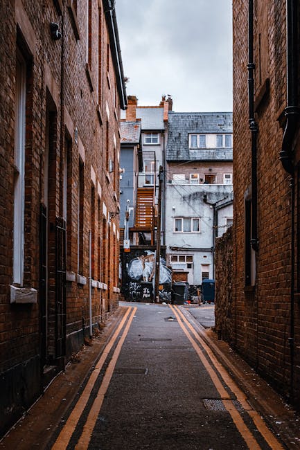 A narrow urban alleyway between two brick buildings, leading to a small open courtyard at the rear. The alley is paved with asphalt and features double yellow lines along both sides, indicating a no-parking zone. On either side, black drainpipes and electrical conduits run vertically along the brick walls, which show signs of weathering and age. At the end of the alley, a wooden staircase with a metal railing ascends to the upper floor of a neighboring building. In the background, several back entrances to residential or commercial properties are visible, including a door and small windows, with some graffiti on a dark wall beneath the staircase. The scene is lit with natural daylight, and the overall setting suggests a typical city environment suitable for home relocation and furniture transport services provided by Man With a Van Abbey Wood.