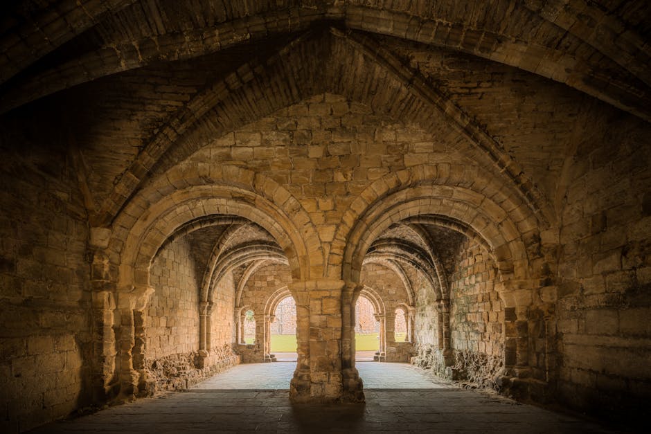 The interior of an ancient stone cloister or arcade with a series of pointed arches supported by thick columns, constructed from weathered sandstone blocks. The vaulted ceiling features ribbed arches, with some areas showing signs of age and erosion. The floor is made of flat stone slabs, and natural light filters in through the small openings at the far end, illuminating the stonework and casting soft shadows. The structure appears to be part of a historical church or abbey, with a view of the outdoor landscape visible through the open arches. This setting exemplifies traditional stonework and architectural craftsmanship, suitable for illustrating house removals or relocation services involving historical or delicate buildings, with [COMPANY_NAME] providing expertise in careful handling and transport of belongings during home or building moves.