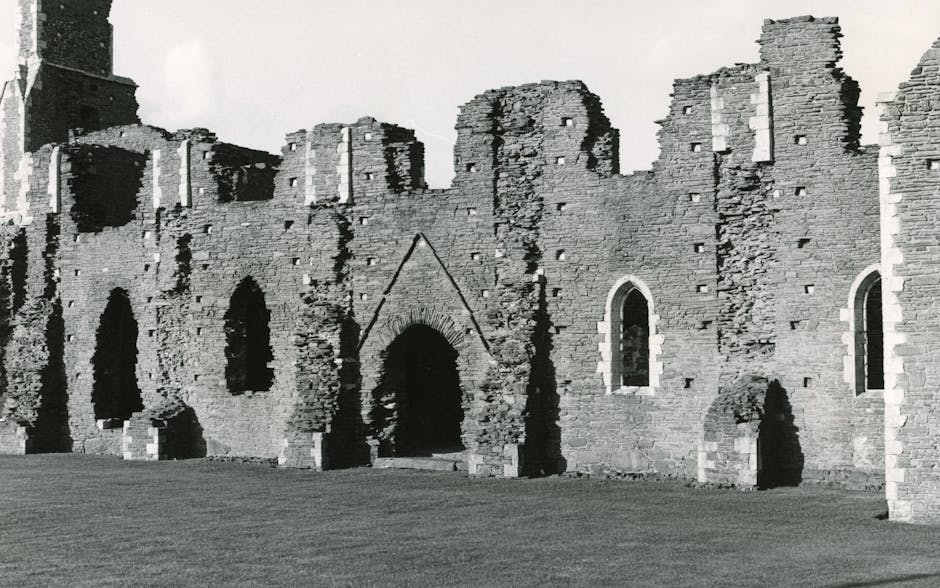A stone ruin of a large historic building, possibly a cathedral or abbey, with multiple arched windows and doorways, some of which are partially collapsed or deteriorated. The structure features tall, pointed Gothic-style arches and vertical stone walls, set on a grassy hill under a cloudy, overcast sky. The ground surrounding the ruin is covered in green grass, with no visible vehicles, furniture, or moving equipment within the scene. This historic site provides a backdrop that could be associated with house removals or relocation services when considering the broader context of moving and transport logistics, as seen on the webpage titled 'Bulky item removal in Abbey Wood: fees & options' from Man With a Van Abbey Wood.