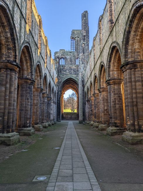 A wide view of the interior of Lesnes Abbey ruins, showing tall, weathered stone walls and arches on both sides, with some sections missing or crumbled. The central walkway is paved with rectangular s