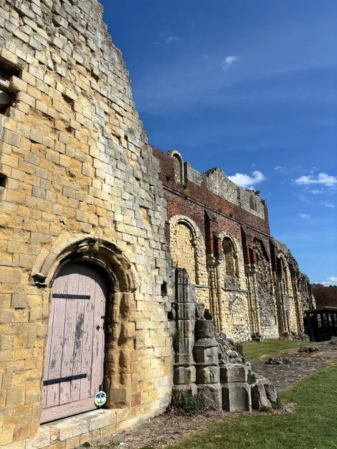 A wide view of the interior of Lesnes Abbey ruins, showing tall, weathered stone walls and arches on both sides, with some sections missing or crumbled. The central walkway is paved with rectangular s