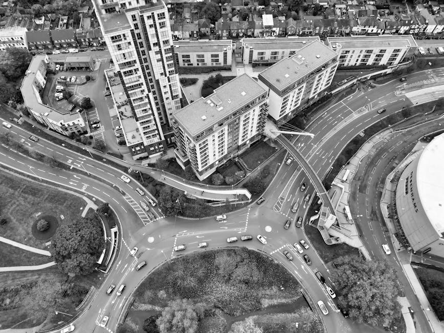 An aerial black and white photograph showing a busy urban area with multiple multi-storey residential buildings, some with balconies, situated along curved roads and a roundabout. Several cars are visible on the streets, some approaching intersections or waiting at traffic lights. A pedestrian bridge connects two of the buildings, and there are wheelchair ramps at street level providing access. The surrounding environment includes landscaped sections with trees and grass patches, indicating an organized cityscape. This scene depicts a typical city centre with high-rise residential housing and road infrastructure, illustrating the logistics involved in house removals and furniture transport services, as offered by Man With a Van Abbey Wood, often necessary during home relocation projects involving packing, loading, and street traffic management.