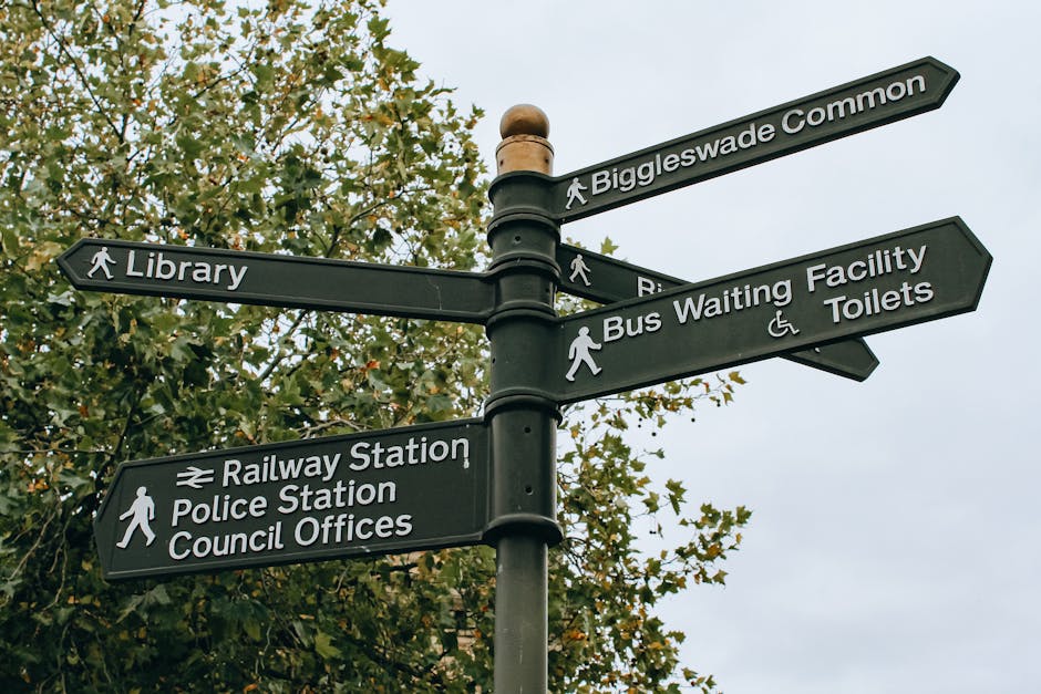 A black multi-directional street signpost located outdoors amidst green foliage, displaying several white text signs with directional arrows. The signs indicate nearby locations including Biggleswade Common, Library, Railways Station, Police Station, Council Offices, Bus Waiting Facility, and Toilets. The signpost is mounted on a tall, rounded pole, with some signs showing walking icons to guide pedestrians. The sky in the background is overcast, casting diffuse natural light. The image captures a typical urban setting that could be relevant for a house or apartment move within Abbey Wood, illustrating the kind of directional signage often encountered during local logistics and planning for furniture transport or home relocation, as part of efficient moving services provided by Man With a Van Abbey Wood.