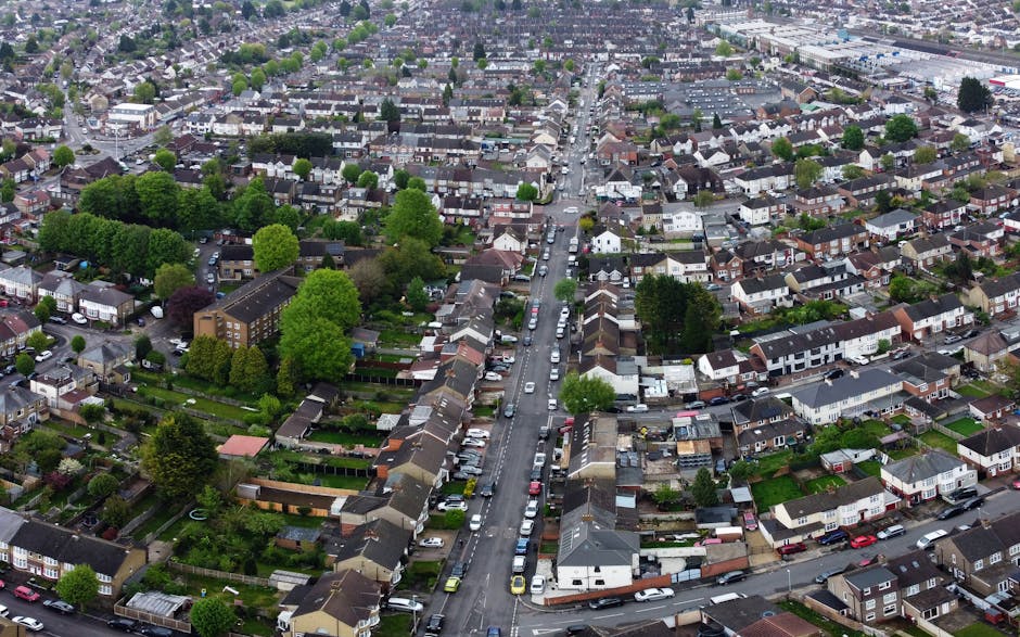 An aerial black and white photograph showing a busy urban area with multiple multi-storey residential buildings, some with balconies, situated along curved roads and a roundabout. Several cars are visible on the streets, some approaching intersections or waiting at traffic lights. A pedestrian bridge connects two of the buildings, and there are wheelchair ramps at street level providing access. The surrounding environment includes landscaped sections with trees and grass patches, indicating an organized cityscape. This scene depicts a typical city centre with high-rise residential housing and road infrastructure, illustrating the logistics involved in house removals and furniture transport services, as offered by Man With a Van Abbey Wood, often necessary during home relocation projects involving packing, loading, and street traffic management.