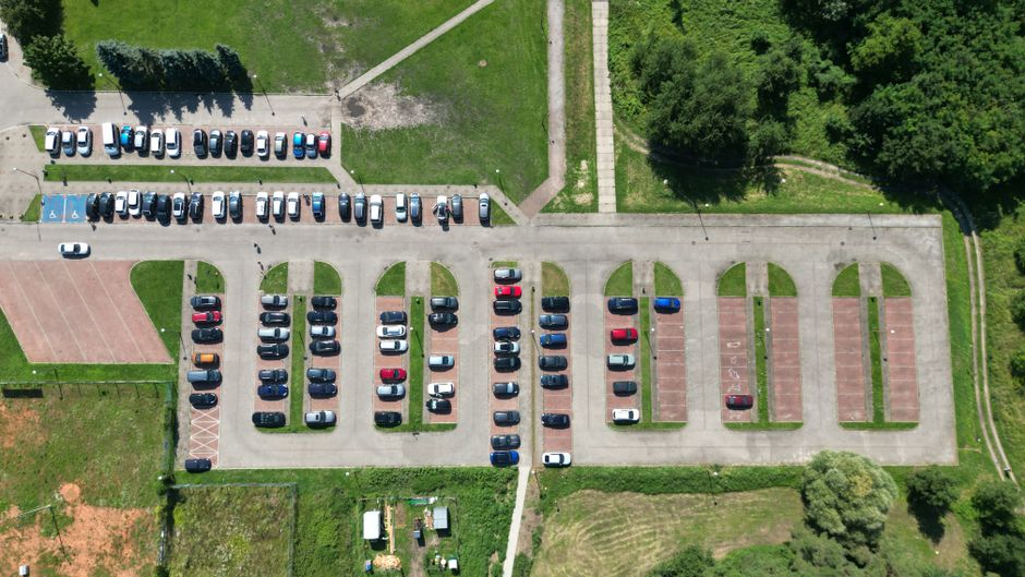 An aerial view of a parking lot adjacent to a park with multiple rows of parked cars in various colours, including white, black, red, and blue. The parking area features designated spaces, some marked for disabled parking, with clear white lines separating each spot. On the left side, there is an outdoor sports court with a reddish-brown surface, and a pathway runs along the top edge of the parking lot, leading into the green park area filled with mature trees and pathways. A man with a van, part of Man With a Van Abbey Wood, is visible in the process of loading or unloading furniture wrapped in plastic and protected with blankets, utilizing a trolley or lift equipment to assist with the home relocation. The van is parked at the edge of the loading zone, with furniture and boxes stacked nearby, ready to be moved into or out of a nearby property. The scene captures the logistics involved in furniture transport and packing and moving activities during house removals, with the environment bathed in daylight.