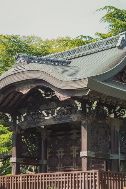 Photograph of a traditional Japanese-style wooden pagoda with ornate roofing, situated outdoors with lush green trees in the background and bright natural lighting. The structure features detailed carved wooden panels and decorative eaves, with the roof extending outward in a curved shape covered in grey tiles. The surrounding environment suggests a tranquil garden or park setting, though no moving objects or furniture are visible. This image does not directly relate to house removals or relocation services, and appears to be of cultural or architectural interest. The image highlights craftsmanship and design typical of Japanese architecture, but does not include any packaging, furniture, or transport equipment related to home relocation or moving processes.