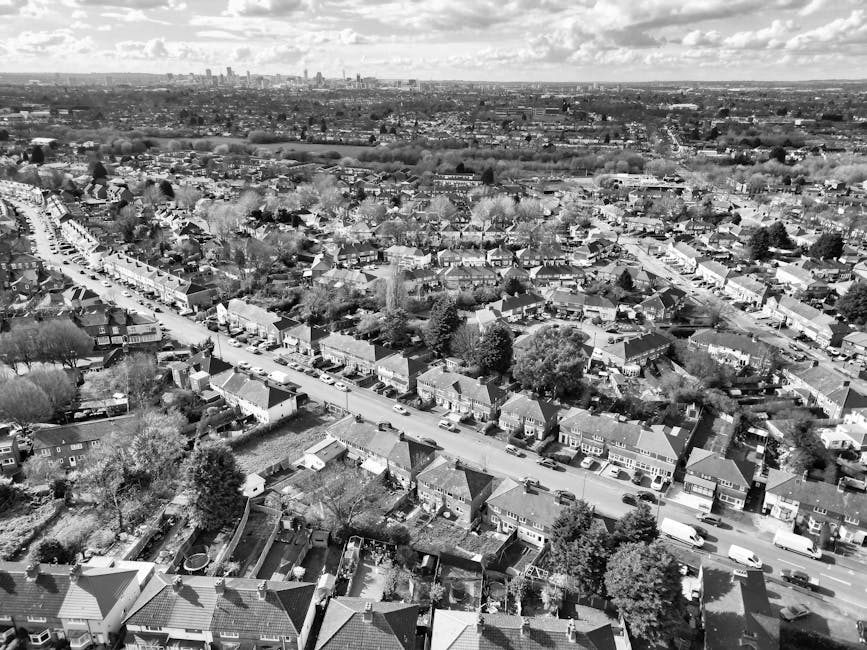 A black-and-white aerial photograph of a residential neighbourhood showing rows of terraced houses and detached homes with pitched roofs, gardens, and trees, extending into the distance with a city skyline visible on the horizon. In the foreground, rooftop details and garden layouts are discernible, with some gardens containing visible fences, sheds, and garden furniture. The streets are lined with parked cars, and the overall scene depicts a typical suburban area with closely packed houses. This image provides context for home relocation or moving services, such as those offered by Man With a Van Abbey Wood, highlighting the typical environment in which furniture transport and packing and moving operations take place within the local community.