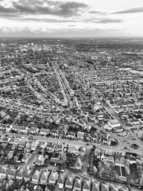 An aerial black-and-white photograph of a suburban residential area with rows of tightly packed houses, each with pitched roofs and small gardens, positioned along a network of streets. In the foreground, a van with open rear doors is visible, parked on a paved driveway adjacent to a house. The van is being loaded with various moving boxes, some wrapped in plastic and others in cardboard, along with pieces of furniture such as a small table and a chair. Moving blankets and straps are used to secure larger items during the loading process. The scene takes place near the entrance of a property, with a clear pathway leading into the home's garden and a sidewalk on the street. The overcast sky above casts soft, diffuse light over the area, emphasizing the detailed layout of the houses and streets. This visual accurately captures a typical house removal process, illustrating the careful packing and loading stage handled by a professional moving service, such as Man With a Van Abbey Wood, during a home relocation effort.