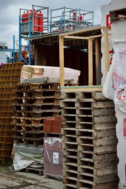 A construction site showing a partially built structure with wooden framework and a metal scaffold on the upper level, which supports multiple red fire extinguishers. In the foreground, there are several stacks of wooden pallets, some carrying wrapped bricks and construction materials, positioned on a paved surface. To the right, a large piece of white construction insulation or sheathing is visible, partially covered by a white protective sheet with red markings. The overall scene depicts ongoing building work, with various materials and equipment arranged around the site, reflecting a typical stage in the home relocation or furniture transport process. The setting is outdoors under cloudy skies, with natural light illuminating the scene, consistent with construction and renovation environments supported by services such as those offered by Man With a Van Abbey Wood.