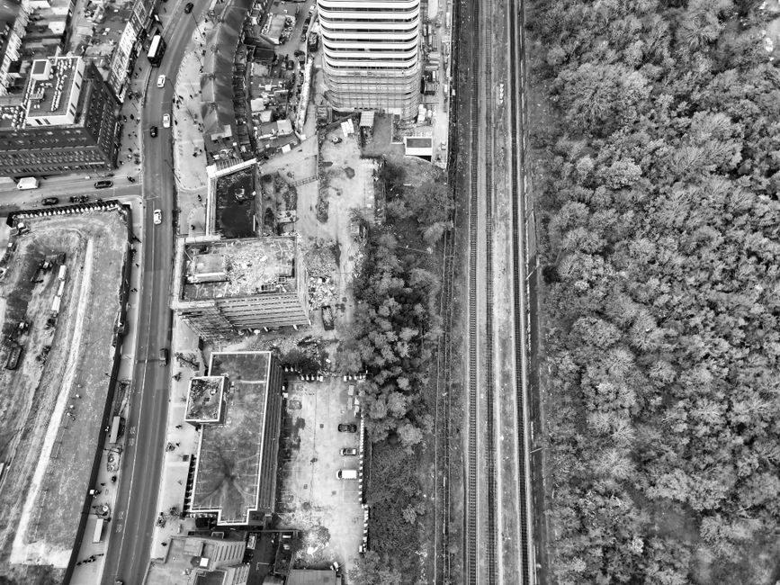 An aerial black-and-white photograph showing an urban environment with a mix of buildings, a railway track running parallel to a densely wooded area on the right, and a paved street with multiple parked and moving vehicles on the left. In the center, there is an open outdoor space with several stacked cardboard boxes, plastic wrapping, and loose packing materials near a partially visible van or truck used for furniture transport and home relocation purposes. The scene depicts a loading or unloading process associated with house removals, with some boxes and materials arranged close to the vehicle, likely being prepared for transport or unpacked after delivery. The lighting appears natural, highlighting the spatial relationship between the urban structures, the railway, and the surrounding greenery, supporting the context of organized removal services provided by Man With a Van Abbey Wood related to packing, loading, and moving logistics near property environments.